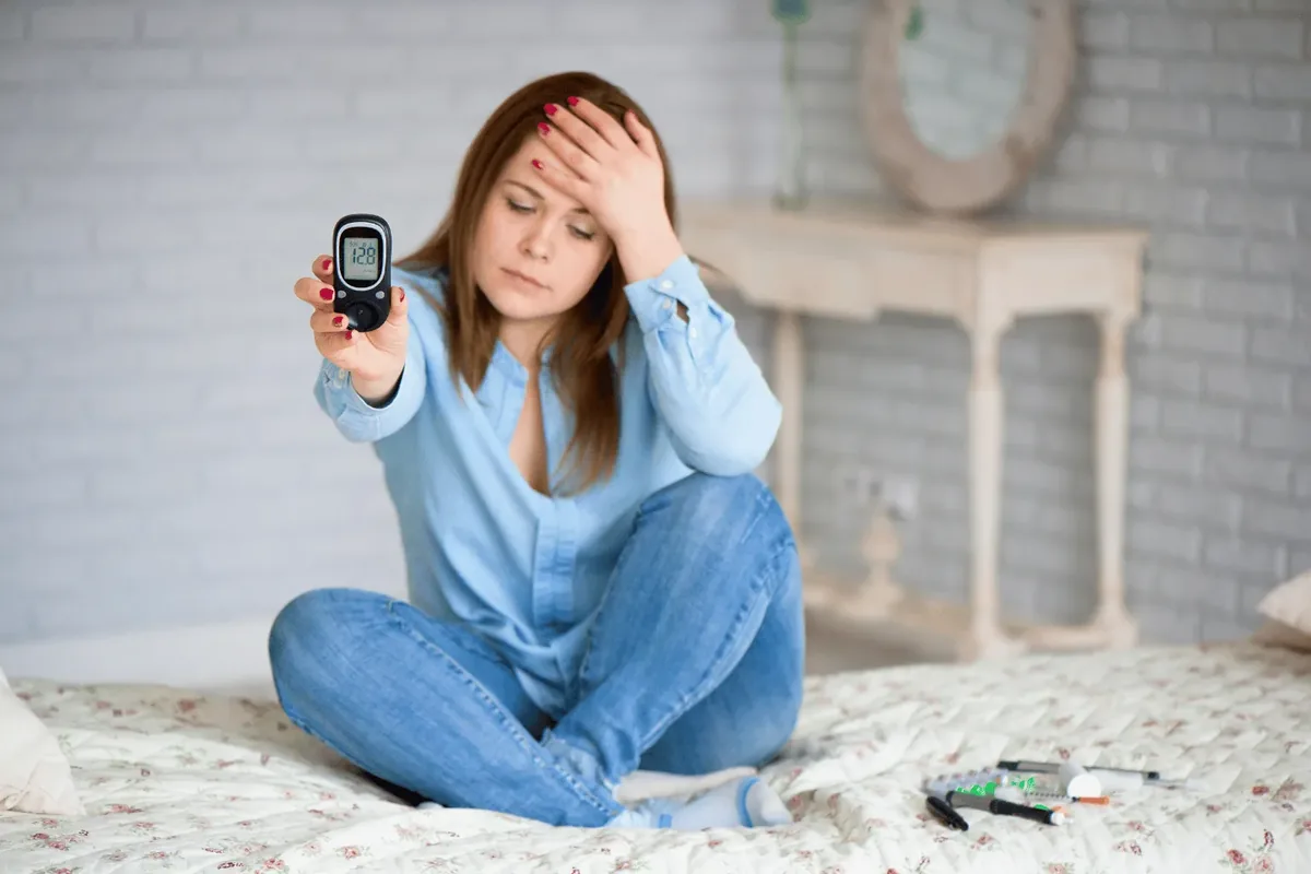 Woman sitting on her bed at home in Dubai reading her blood glucose level on a handheld glucose meter, with lancets and insulin pens beside her
