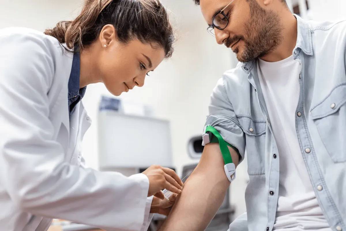 A clinician in a white coat drawing a venous blood sample from a seated patient’s arm with a tourniquet in place — the single blood draw a lipid and cholesterol profile test requires