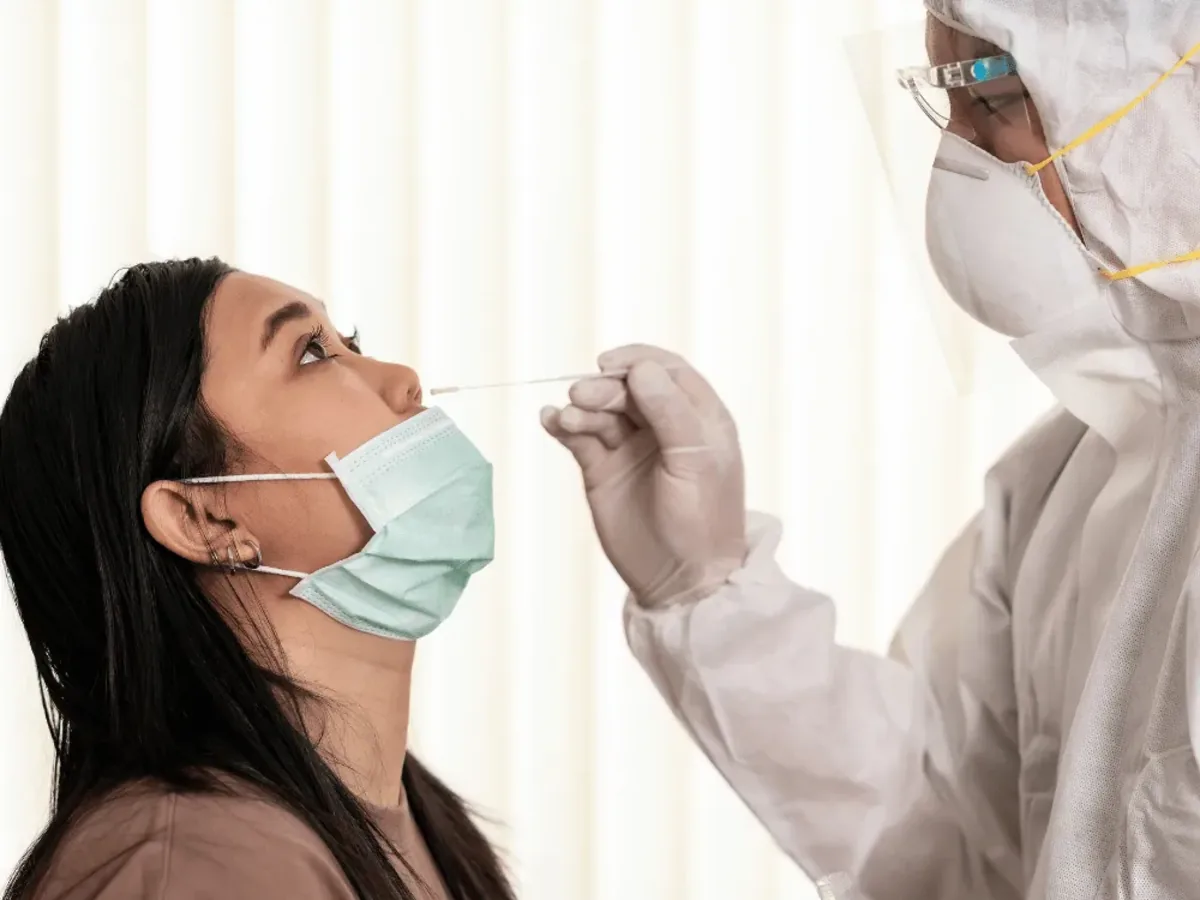 A healthcare worker in full protective equipment and a face shield collecting a nasal swab sample from a masked patient for a Covid 19 PCR test at home in Dubai