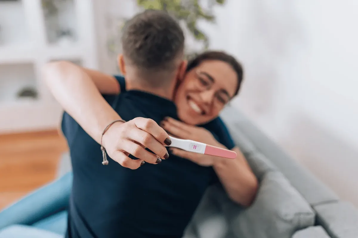 A smiling woman embracing her partner at home while holding a positive pregnancy test, illustrating a pregnancy test taken at home in Dubai